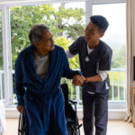 Diverse male doctor helping senior male patient getting up from wheelchair at home. Medical services, check up, home visit, healthcare, wellbeing and senior lifestyle, unaltered.