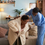Senior Black woman using cane receiving assistance from young adult Black female nurse in scrubs helping her stand up from sofa in living room setting during home care visit