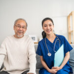 Portrait of Asian caregiver nurse sit on sofa and take care senior man. Attractive specialist carer women support older elderly mature grandfather at nursing home care and smiling, looking at camera.