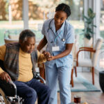 African American senior man getting up from his wheelchair with help of young nurse at residential care home.