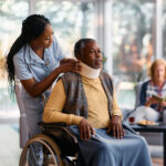 African American nurse adjusting neck collar to elderly man who is sitting in wheelchair at nursing home.
